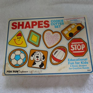 May include: A box of Fox Run Craftsmen cookie cutters in the shape of a triangle, square, heart, school bus, stop sign, soccer ball, dog, and football. The box says "Shapes Cookie Cutter Set" and "Educational Fun for Kids 8 Basic Shapes for Creative Hands".
