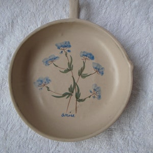 May include: A beige ceramic serving dish with a handle, featuring a hand-painted floral design in blue and green. The word "anise" is written in blue script below the flowers. The dish has a round shape.