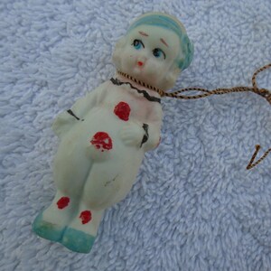 May include: A vintage porcelain doll figurine in a clown-like outfit. The doll has a white body with red polka dots, blue shoes, and a blue cap. A brown string is attached to the doll's neck.