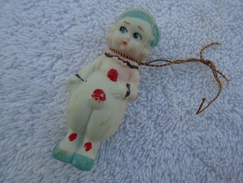 May include: A vintage porcelain doll figurine in a clown-like outfit. The doll has a white body with red polka dots, blue shoes, and a blue cap. A brown string is attached to the doll's neck.