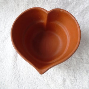 May include: A heart-shaped, terracotta-colored bowl. The bowl has a smooth interior and a slightly rough exterior. The heart shape is clearly defined, with rounded lobes and a pointed base. The bowl is empty and sits on a white, textured surface.