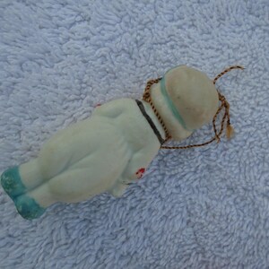 May include: A vintage porcelain doll in a sailor outfit, lying on its back. The doll is primarily white with blue accents on the shoes and hat. It has a black collar and a brown string for hanging.