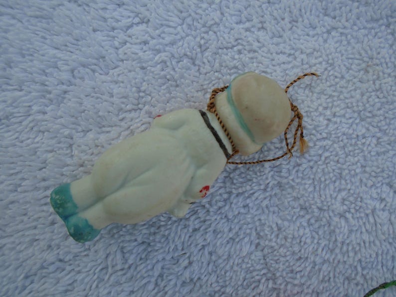 May include: A vintage porcelain doll in a sailor outfit, lying on its back. The doll is primarily white with blue accents on the shoes and hat. It has a black collar and a brown string for hanging.