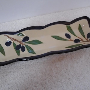 May include: A rectangular ceramic serving dish with a wavy edge and a cream-colored interior. The dish features a hand-painted design of brown branches with dark blue olives and green leaves. The rim is finished with a thick, dark brown border.