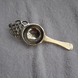 May include: A silver-toned tea infuser with a round, perforated bowl and a long handle. The infuser has an ornate, decorative design at the top, and is designed for steeping loose leaf tea. The infuser is shown on a gray surface.