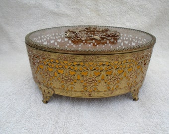 May include: A gold-toned, oval-shaped jewelry box with a glass lid and intricate floral design. The box has a raised floral design on the lid and sits on four small feet.