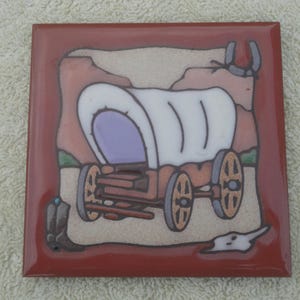 May include: Square ceramic tile with a painted image of a covered wagon, a cowboy boot, and a horseshoe. The wagon has a white cover and purple interior. The tile has a red border and a beige background.