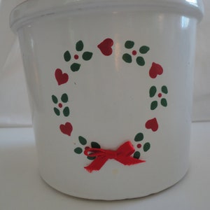 May include: White ceramic crock with a decorative wreath of red hearts and green leaves. A red bow is tied at the bottom of the wreath. The crock has a slightly flared shape and a rounded rim. The design is hand-painted.