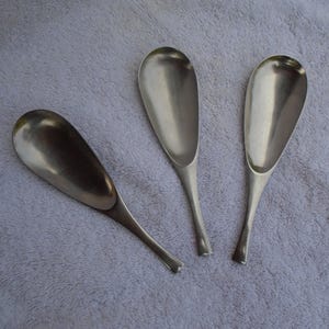 May include: Three silver-colored scoops with elongated, oval bowls and slender handles are arranged on a textured, light gray surface. The scoops are made of a shiny metal, reflecting light.