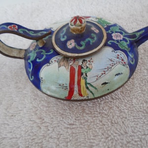 May include: A small, round teapot with a blue enamel finish and a decorative lid. The teapot features a handle and spout, and is adorned with floral and landscape designs in various colors. The lid has a red and white floral design.