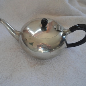 May include: A silver teapot with a black handle and a black knob on the lid. The teapot is sitting on a white fluffy surface.