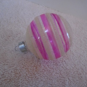 May include: A round, clear glass Christmas ornament with diagonal pink stripes. The ornament has a silver metal cap and hook for hanging. The stripes are a vibrant pink color, and the ornament has a glittery finish.