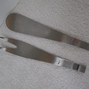 May include: Two stainless steel serving utensils: a long, flat spatula and a two-pronged fork. The spatula has a rounded end, while the fork has sharp, pointed tines. Both are lying on a textured, light gray surface.