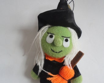 May include: A plush Halloween witch doll with a green face, white hair, and a black hat and dress. The doll has an orange bow and holds an orange broom. A black string is attached to the hat.