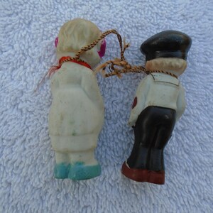 May include: Two small vintage figurines, a sailor and a doll, connected by a string. The sailor wears a black hat, white shirt, and black pants. The doll has a white coat, pink eyes, and blue shoes. The figures are made of a light-colored material.