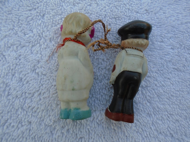 May include: Two small vintage figurines, a sailor and a doll, connected by a string. The sailor wears a black hat, white shirt, and black pants. The doll has a white coat, pink eyes, and blue shoes. The figures are made of a light-colored material.