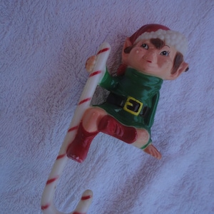 May include: A vintage Christmas stocking holder featuring a green and red elf holding a candy cane. The elf has a white hat with a red trim and a brown face with a white beard.