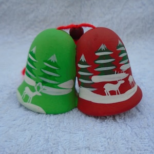 May include: Two ceramic Christmas bells, one green and one red, with white painted winter scenes of trees and deer. The bells are connected with a red string and a small wooden bead.