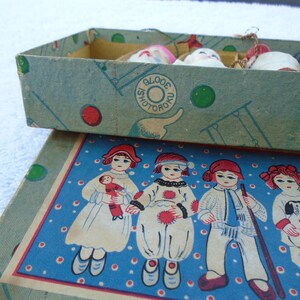 May include: Vintage box with a blue and green design, containing small doll heads. The box lid features illustrations of children in various outfits on a blue background. The box has the text "SHOTORO".