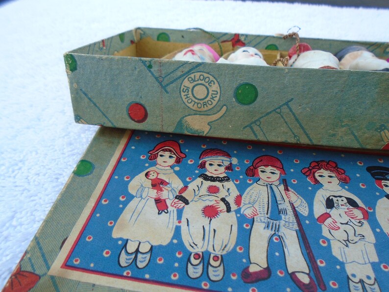 May include: Vintage box with a blue and green design, containing small doll heads. The box lid features illustrations of children in various outfits on a blue background. The box has the text "SHOTORO".
