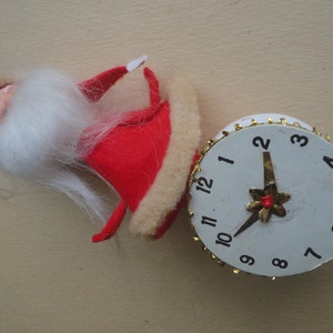 May include: A vintage Christmas ornament featuring a red Santa Claus figure with a white beard and a white clock with gold trim. The clock hands point to 10:58. The ornament is on a beige surface.