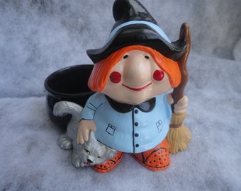 May include: A ceramic witch figurine with a black hat and broom, wearing a light blue shirt and orange shoes. A black cauldron and a gray cat are also present. The witch has red hair and rosy cheeks. A Halloween-themed decoration.