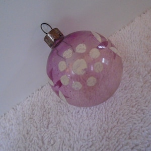 May include: A vintage Christmas ornament. The glass sphere is a soft pink with white polka dots and floral accents. It has a metal cap and a wire loop for hanging. The ornament is resting on a white, textured surface.