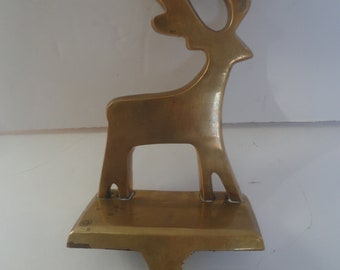 May include: A brass-colored reindeer stocking holder. The reindeer silhouette has detailed antlers and stands on a rectangular base with a hook for hanging. The metal has a warm, golden hue.
