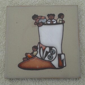 May include: Square ceramic tile featuring a stylized illustration of a white boot with brown and orange accents. Several small figures are depicted, some sitting and others peering over the top of the boot. The tile has a light beige background.