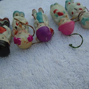 May include: A collection of vintage ceramic doll ornaments. The ornaments feature painted faces, colorful clothing, and attached string loops for hanging. The dolls have various outfits, including hats and dresses, and are arranged on a white surface.