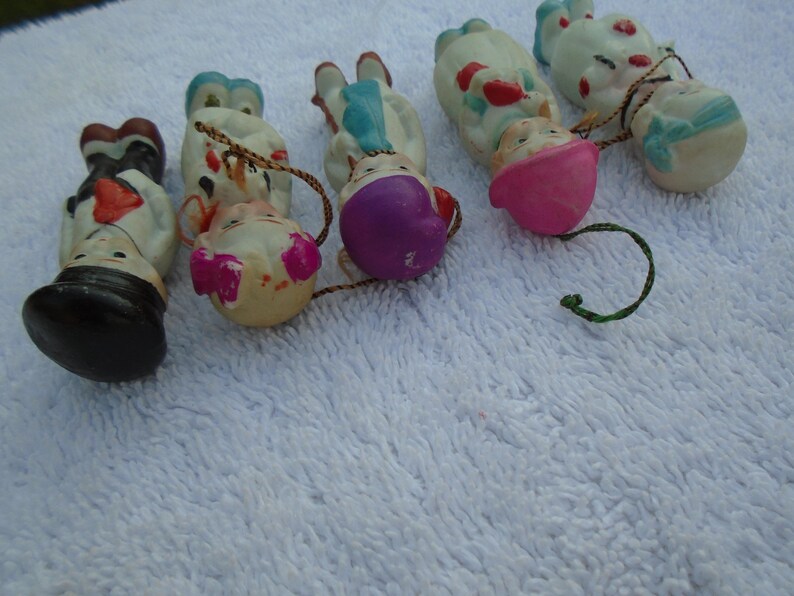 May include: A collection of vintage ceramic doll ornaments. The ornaments feature painted faces, colorful clothing, and attached string loops for hanging. The dolls have various outfits, including hats and dresses, and are arranged on a white surface.