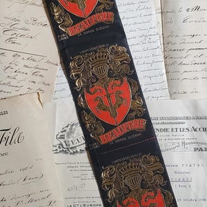 May include: A long, black fabric strip featuring three repeating gold and red crests. Each crest displays a shield with two horses, a sword, and a crown. The text "BEAUFORT" is printed in red, with "IMPERMEABLE" and "PURE LAINE" in gold.
