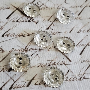 May include: A set of six small, clear, circular buttons with a scalloped edge and two central holes. Each button features a faceted, reflective surface that catches the light, creating a bright, transparent appearance against the background.