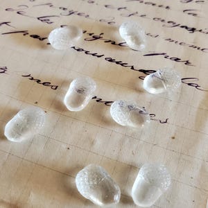 May include: Seven clear glass acorn-shaped beads with a textured cap and a smooth body. Each bead features a distinct, faceted top section resembling the cap of an acorn, while the lower portion is smooth and transparent, reflecting light clearly.