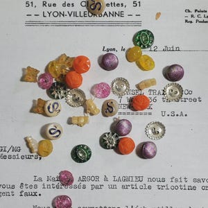 May include: A collection of assorted small buttons in various shapes, colors, and materials, including round orange, purple, green, and clear buttons, some with metallic finishes, and white buttons featuring a blue snake graphic, scattered across a document.