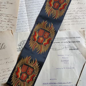 May include: A long, dark blue fabric strip featuring three repeating embroidered crests. Each crest displays a central shield with red and gold details, surrounded by golden flame-like motifs and the text "FOULARD BASQUE" and "IMPERMEABLE".