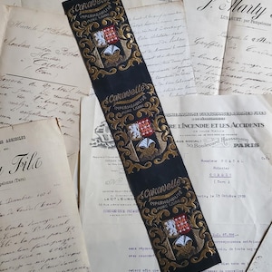 May include: A long, black fabric strip featuring three repeating gold embroidered crests and the text Caravelle Impermeabilise Pure Laine. The strip is layered over a collection of vintage, handwritten and printed French documents with cursive ink.