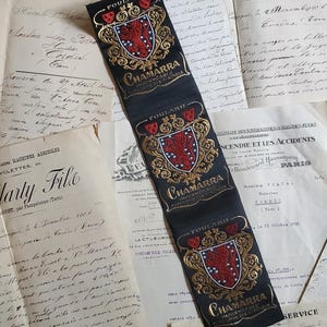 May include: A long black fabric strip featuring three repeating embroidered crests in gold and red. Each crest displays a central red shield with a lion, surrounded by gold filigree. The text FOULARD and CHAMARRA IMPERMEABLE are stitched in gold thread.