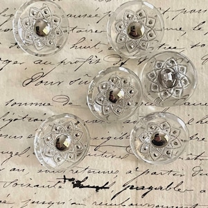 May include: Six circular clear glass buttons, each featuring a central metallic silver-toned dome surrounded by a detailed, star-shaped filigree pattern with small raised dots, arranged in a cluster on a surface with handwritten cursive script.