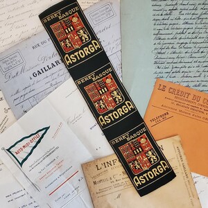 May include: A black fabric ribbon featuring the repeated text BERET BASQUE ASTORGA in gold lettering, accompanied by a red and gold heraldic crest. The ribbon is layered over a collection of vintage off-white and orange paper documents with text.