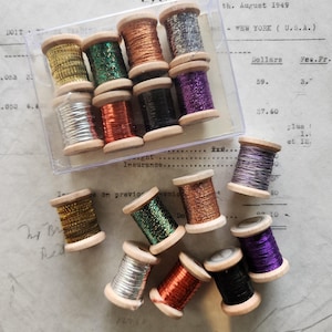 May include: A collection of metallic embroidery thread spools in various colors including gold, silver, copper, green, purple, and black. Eight spools are housed in a clear plastic box, while seven additional spools are arranged outside the box.