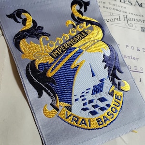 May include: A light gray fabric strip featuring two identical embroidered crests in blue, yellow, and white. Each crest displays a shield with a building, surrounded by ornate scrollwork and the text "Kossoc" and "IMPERMEABLE VRAI BASQUE".