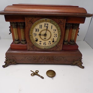 May include: A wooden antique mantel clock with a round clock face, Roman numerals, and decorative columns. The clock has a key and a pendulum weight. The clock is brown with gold accents and ornate details.