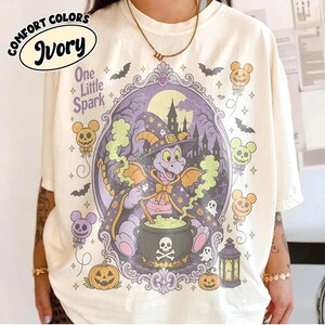 Puede incluir: Una camiseta color marfil con un estampado gráfico de un dragón morado con sombrero de bruja y capa, revolviendo un caldero negro con un diseño de calavera. La escena incluye un castillo encantado, murciélagos, fantasmas y globos de calabaza.