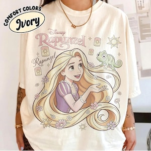 May include: An ivory t-shirt featuring a graphic of Rapunzel with long blonde hair, a purple dress, and a green chameleon. The design includes floating lanterns, small flowers, a sun icon, and the text Disney Rapunzel. A Comfort Colors Ivory label is visible.