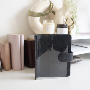 Può includere: Un planner in pelle nera con una copertina in plastica trasparente e una patta in pelle nera con chiusura a bottone.