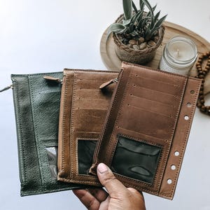 May include: Three leather wallets in shades of green and brown. The wallets have a zippered closure, clear ID windows, and multiple card slots. One wallet is open, revealing the interior design. The wallets are designed for organizing cards and cash.