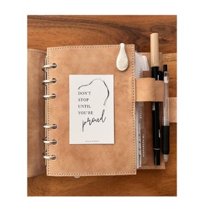 May include: A tan leather bound planner with a white card that says "Don't stop until you're proud." The card is in the left side of the planner. The right side of the planner has a pen holder with three pens. The planner is on a wooden surface.