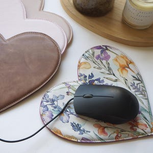 Könnte beinhalten: Herzförmige Mauspads aus rosa und braunem Leder. Ein Pad zeigt ein Blumenmuster mit lila, orangefarbenen und grünen Blumen auf weißem Grund. Eine schwarze Computermaus liegt auf dem floralen Pad.