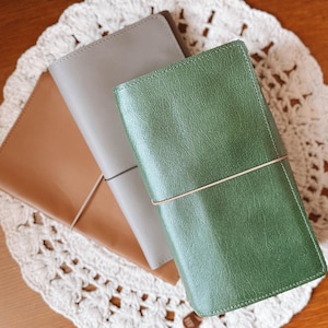 May include: Three leather journals in shades of brown, gray, and green are displayed on a white, crocheted doily. Each journal is rectangular with a simple elastic closure. The journals are a neutral accessory.